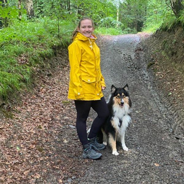 Frau in einer gelben Jacke steht auf einem Weg in einem Park mit einem Hund und schaut in die Kamera.