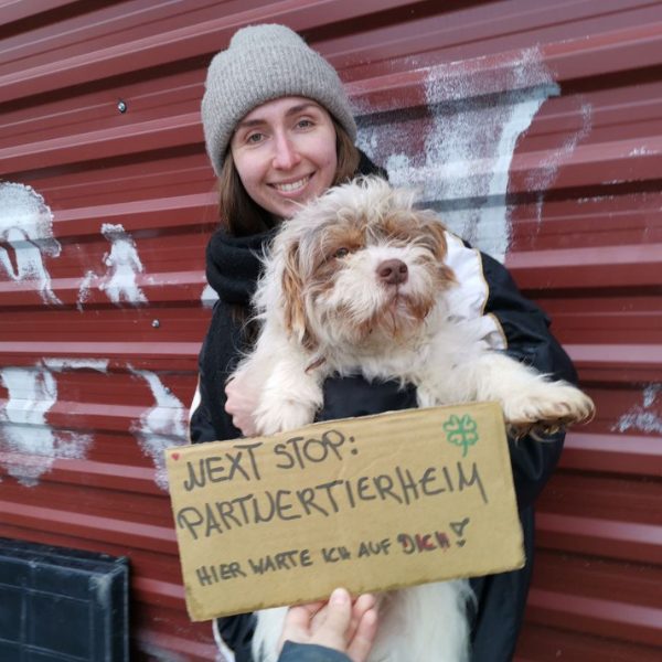 Frau mit Hunde auf dem Arm und Schild schaut in die Kamera
