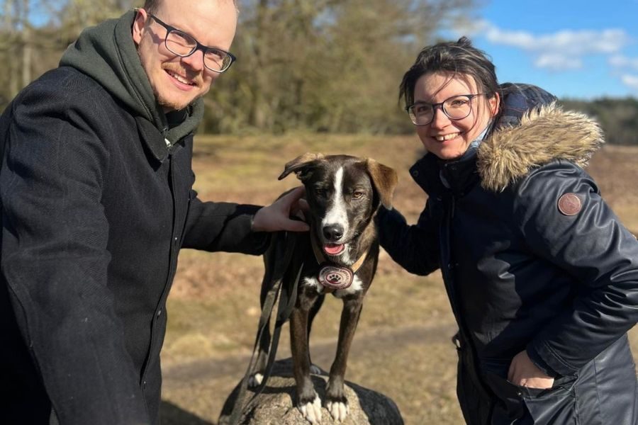 Zwei Menschen haben einen Hund in ihrer Mitte und schauen in die Kamera.