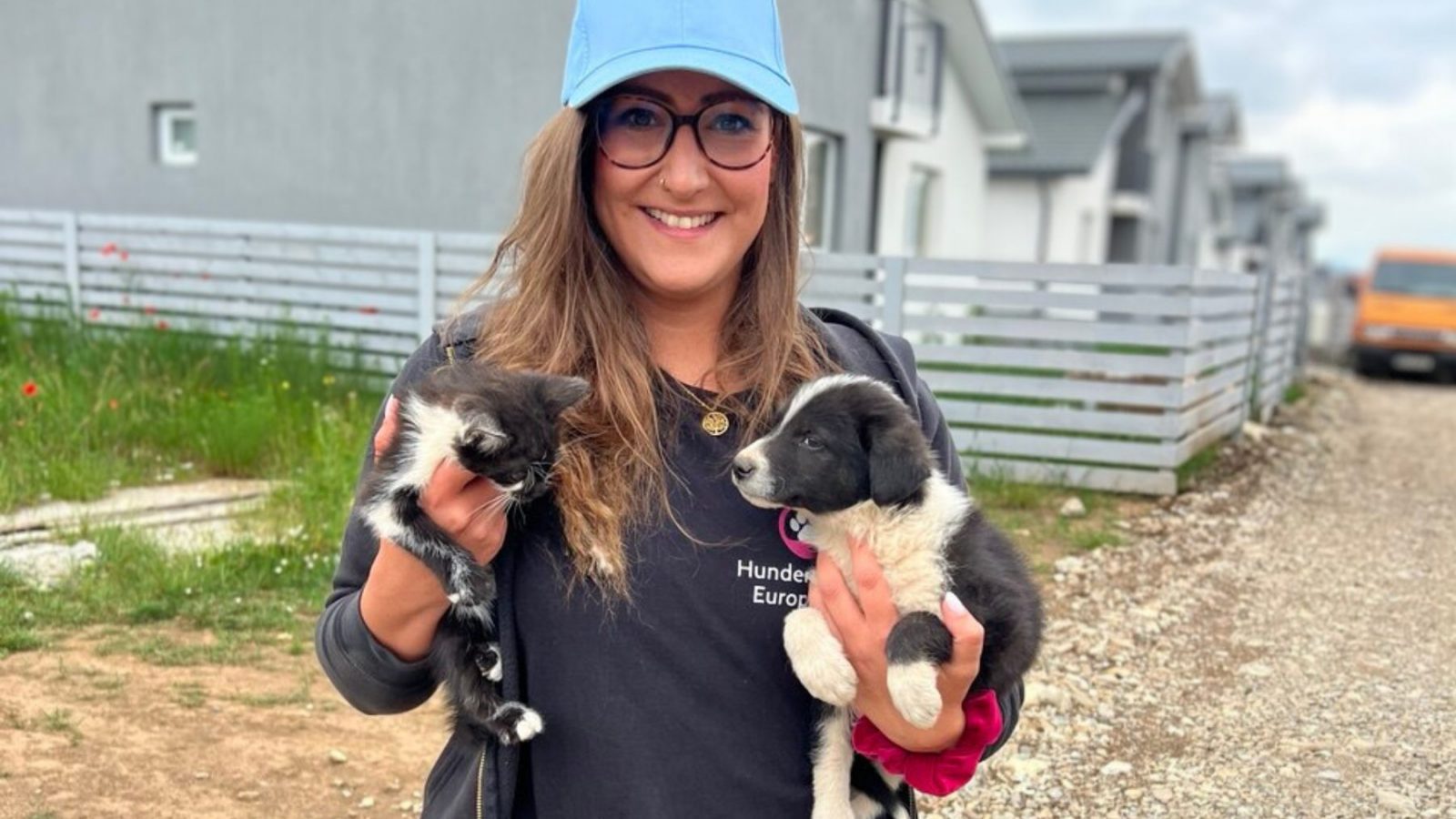 Frau mit blauem Cap hält in der linken Hand ein schwarz-weißes Katzenbaby und in der rechten Hand ein schwarz-weißes Hundebaby.