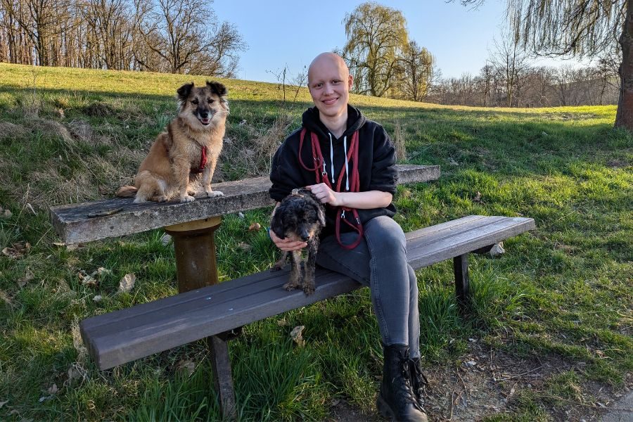 Eine Frau sitzt mit ihren zwei kleinen Hunden auf einer Bank und lächelt.