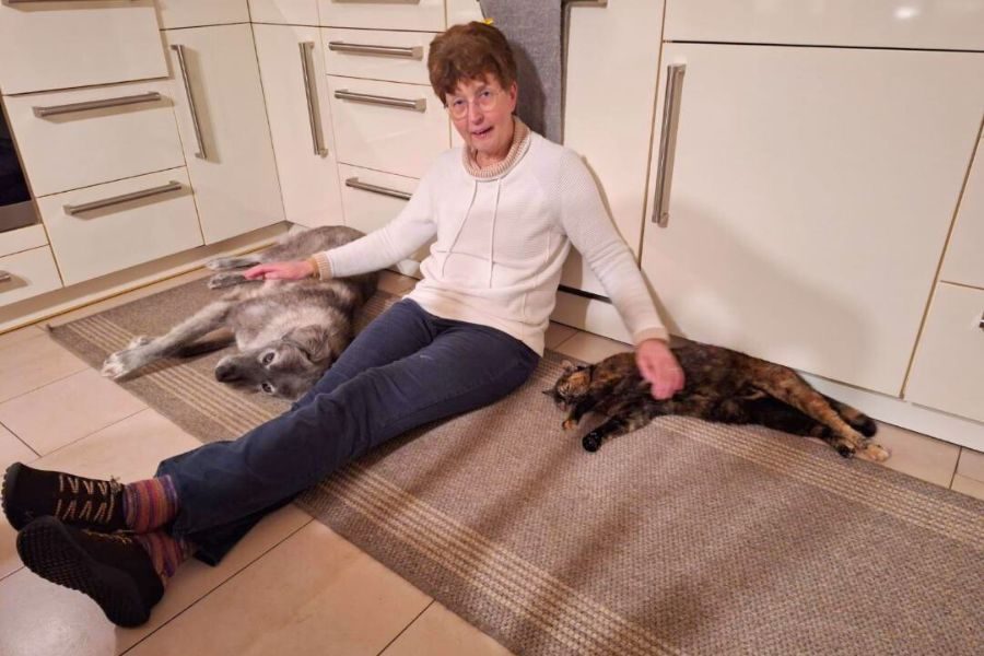 Eine Frau sitzt mit einem Hund und einer Katze auf dem Fußboden und streichelt die Tiere.